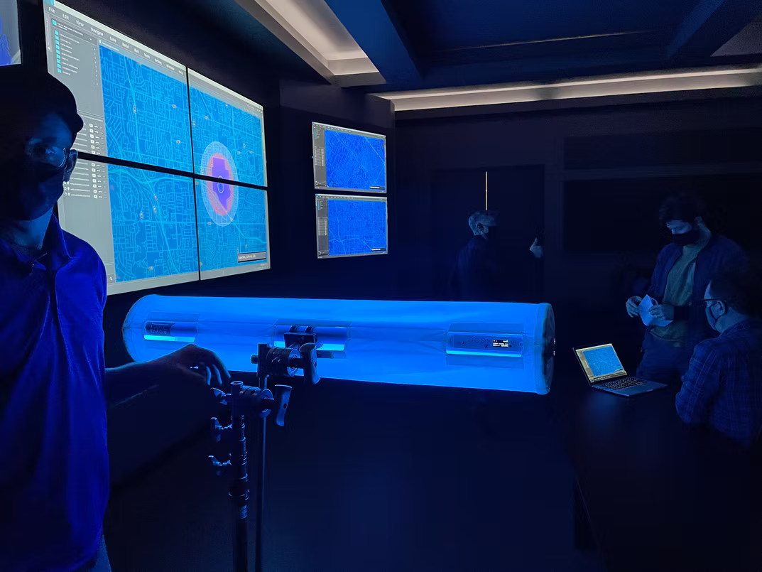 A large cylindrical device glowing with bright blue light sits on a table in a dark room, surrounded by several people wearing face masks. Multiple large screens on the wall display blue data visualizations, and a camera on a tripod is positioned in front of the device.